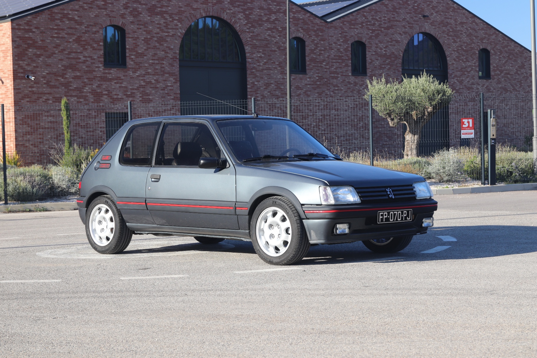 Peugeot 205 GTI 1.9 130 ch gris graphite de profil sur parking devant bâtiment en briques